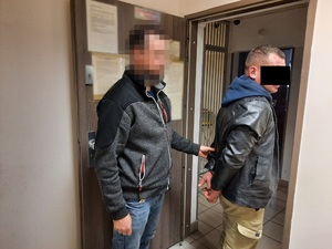 Zdjęcie przedstawia policjanta ubranego w ciemnoszarą bluzę z czarnymi wstawkami i niebieskie spodnie jeansowe, który stoi za zatrzymanym mężczyzną. Twarz policjanta jest zamazana komputerowym filtrem. Mężczyzna ubrany jest w czarną kurtkę spod której wystaje niebieski kaptur i beżowe spodnie materiałowe. Część twarzy zasłania mu czarny prostokąt. Na rękach trzymanych z tyłu ma założone kajdanki. Obaj znajdują się w pomieszczeniu dla osób zatrzymanych.