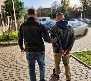 Zdjęcie przedstawia policjanta ubranego w ciemnoszarą bluzę z czarnymi wstawkami i niebieskie spodnie jeansowe, który prowadzi po swojej prawej stronie zatrzymanego mężczyznę. Twarz policjanta jest zamazana komputerowym filtrem. Mężczyzna ubrany jest w czarną kurtkę spod której wystaje niebieski kaptur i beżowe spodnie materiałowe. Na rękach trzymanych z tyłu ma założone kajdanki. Obaj idą po dziedzińcu komendy. W tle widać zaparkowane pojazdy różnych marek.