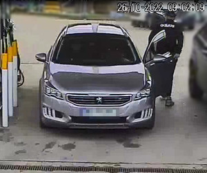 Zdjęcie przedstawia mężczyznę stojącego pomiędzy samochodem - szarym peugeotem, a innym ciemnym zaparkowanym po jego prawej stronie. Mężczyzna ma około 180 cm wzrostu, szczupłą budowę ciała; ubrany jest w czarną czapkę z daszkiem, czarną bluzę z białym logiem ,,Adidas” na plecach, czarne spodnie dresowe, szare obuwie sportowe z białą podeszwą. Samochód zaparkowany jest przodem do kamery.