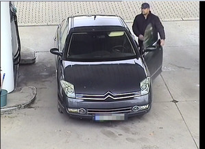 Zdjęcie przedstawia mężczyznę, który ma około 175 cm wzrostu; ubrany jest w czarną czapkę z daszkiem, czarną kurtkę, szare spodnie dresowe i białe obuwie sportowe z czarnymi paskami z logo ,,Adidas”. Stoi przy zaparkowanym ciemnym citroenie. Samochód zaparkowany jest przodem do kamery monitoringu.
