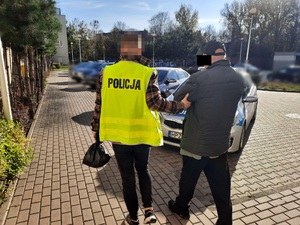 Zdjęcie przedstawia policjanta ubranego w seledynową kamizelkę odblaskową z napisem POLICJA na plecach, który prowadzi po dziedzińcu, po swojej prawej stronie zatrzymanego mężczyznę. Twarz policjanta jest zasłonięta. Mężczyzna jest ubrany na ciemno. Na głowie ma czarną czapkę z daszkiem. Część jego twarzy jest zasłonięta. W tle widać zaparkowany oznakowany radiowóz i inne cywilne samochody.