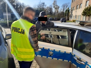 Zdjęcie przedstawia policjanta ubranego w seledynową kamizelkę odblaskową z napisem POLICJA na plecach, który stoi przy oznakowanym radiowozie. Twarz policjanta jest zasłonięta. Policjant stoi tyłem do zdjęcia. Trzyma otwarte drzwi tylne, w których stoi zatrzymany mężczyzna.  Mężczyzna jest ubrany na ciemno. Na głowie ma czarną czapkę z daszkiem. Część jego twarzy jest zasłonięta czarnym prostokątem. W tle widać zaparkowany oznakowany radiowóz i inne cywilne samochody.