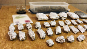 Zdjęcie przedstawia leżące na brązowym stole foliowe zawiniątka, pojemnik plastikowy z suszem roślinnym i słoiczek z suszem.