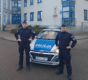Zdjęcie przedstawia dwóch umundurowanych policjantów, którzy stoją po obu stronach oznakowanego radiowozu i patrzą wprost d aparatu. W tle widać budynek komisariatu.
