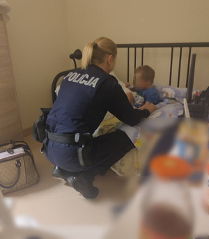 Zdjęcie przedstawia umundurowaną policjantkę, która kuca przy łóżku, na którym siedzi mały chłopczyk. Twarz dziecka jest zasłonięta komputerowym filtrem.