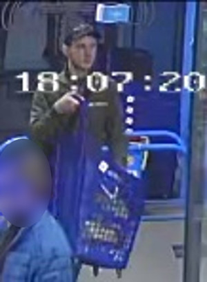 Zdjęcie przedstawia mężczyznę wewnątrz sklepu. Jest on w wieku około 30 lat, ma około 180 cm wzrostu; ubrany jest w zieloną kurtkę, czarną czapkę z daszkiem. Przed sobą, na wysokości klatki piersiowej trzyma niebieski wózek sklepowy. Mężczyzna skierowany jest przodem do kamery monitoringu.