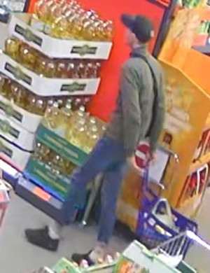 Zdjęcie przedstawia mężczyznę wewnątrz sklepu. Mężczyzna ma około 30 lat, około 180 cm wzrostu; ubrany jest w zieloną kurtkę, niebieskie spodnie, czarne buty sportowe i czarną czapkę z daszkiem, przez ramię ma przewieszoną czarną saszetkę. Za sobą prowadzi niebieski wózek sklepowy. Mężczyzna jest ustawiony lewym profilem do kamery. W tle widać półki z asortymentem.