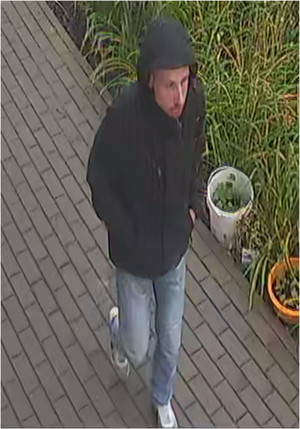 Zdjęcie przedstawia idącego chodnikiem mężczyznę. Ma on  około 30-35 lat, posiada zarost w postaci wąsów; ubrany jest w czarną kurtkę z założonym na głowę kapturem, spodnie jeansowe, białe buty sportowe. Ręce trzyma w kieszeniach kurtki. Znajduje się przodem do obiektywu kamery.