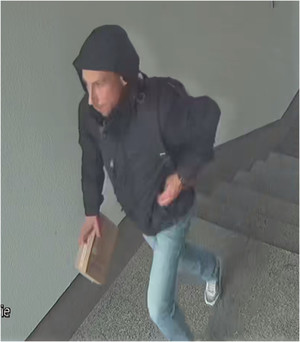 Zdjęcie przedstawia wchodzącego po schodach mężczyznę. Ma on około 30-35 lat, posiada zarost w postaci wąsów; ubrany jest w czarną kurtkę z założonym na głowę kapturem, spodnie jeansowe, białe buty sportowe z czarnymi elementami. W prawe ręce trzyma brązowe kartonowe pudełko. Znajduje się lewym profilem do obiektywu kamery.