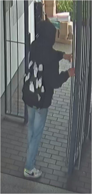 Zdjęcie przedstawia wychodzącego przez okratowaną furtkę mężczyznę. Ubrany jest w czarną kurtkę z założonym na głowę kapturem, spodnie jeansowe, białe buty sportowe z czarnymi elementami. Posiada czarny plecak z białymi elementami. Stoi tyłem do obiektywu kamery.