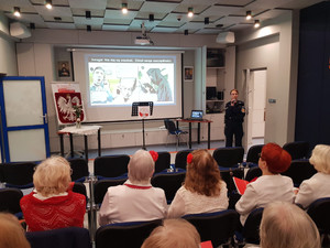Zdjęcie przedstawia umundurowaną policjantkę, która stoi przodem do siedzących na krzesłach seniorów. Za policjantką jest wyświetlony slajd prezentacji dot. oszustw.