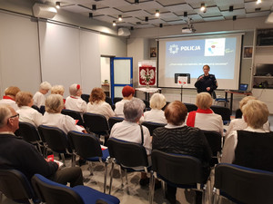 Zdjęcie przedstawia umundurowaną policjantkę, która stoi przodem do siedzących na krzesłach seniorów. Za policjantką jest wyświetlony slajd prezentacji dot. oszustw.