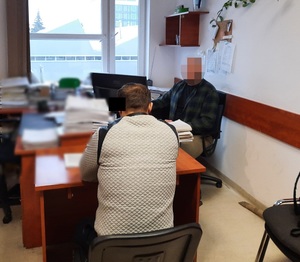 Zdjęcie przedstawia zatrzymanego mężczyznę, który siedzi przy biurku tyłem do wejścia do pokoju. Po jego prawej stronie siedzi nieumundurowany policjant. Jego twarz jest zasłonięta.