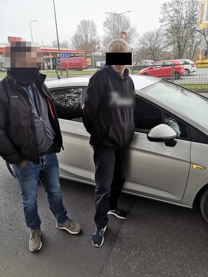 Zdjęcie przedstawia nieumundurowanego policjanta, który stoi z zatrzymanym mężczyzną przy srebrnym samochodzie. Zatrzymany mężczyzna jest ubrany na ciemno, część twarzy zasłana mu czarny prostokąt. Twarz policjanta jest rozmazana komputerowym filtrem.