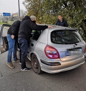 Zdjęcie przedstawia nieumundurowanych policjantów, którzy stoją po obu stronach srebrnego pojazdu. Zatrzymany mężczyzna trzyma dłonie na dachu samochodu. Zatrzymany mężczyzna jest ubrany w czarną kurtkę i ciemne spodnie, część twarzy zasłana mu czarny prostokąt. Twarze policjantów są rozmazane komputerowym filtrem.