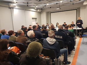 Zdjęcie przedstawia policjantów, strażników miejskich i innych przedstawicieli instytucji siedzących za długim stołem naprzeciwko ludzi siedzących na sali. Wśród tych ludzi siedzą umundurowani policjanci.