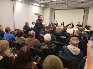 Zdjęcie przedstawia policjantów, strażników miejskich i innych przedstawicieli instytucji siedzących za długim stołem naprzeciwko ludzi siedzących na sali. Wśród tych ludzi siedzą umundurowani policjanci. Jeden z policjantów stoi przed zebranymi.