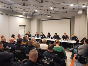 Zdjęcie przedstawia policjantów, strażników miejskich i innych przedstawicieli instytucji siedzących za długim stołem naprzeciwko ludzi siedzących na sali. Umundurowana policjantka stoi przed zebranymi.