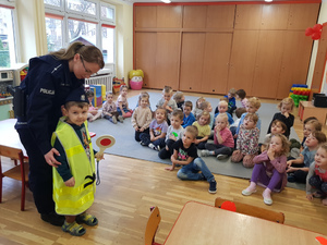 Zdjęcie przedstawia umundurowaną policjantkę, która stoi obok dziecka ubranego w policyjną odblaskową kamizelkę i czapkę służbową, przed nimi na dywanie siedzą inne dzieci.