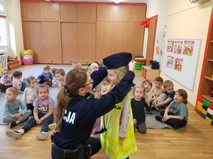 Zdjęcie przedstawia umundurowaną policjantkę, która kuca obok dziecka ubranego w policyjną odblaskową kamizelkę i czapkę służbową, za nimi na dywanie siedzą inne dzieci.