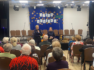 Zdjęcie przedstawia dwie umundurowane policjantki, które soją przed siedzącymi na krzesłach seniorami.