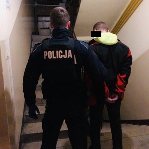 Zdjęcie przedstawia umundurowanego policjanta, który prowadzi po klatce schodowej zatrzymanego mężczyznę. Mężczyzna ten ma kajdanki założone na rękach trzymanych z tyłu. Jest ubrany w czarną kurtkę z czerwonymi wstawkami, spod której wystaje jasny kaptur bluzy.