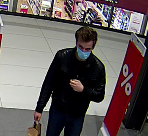 Zdjęcie przedstawia mężczyznę wchodzącego do sklepu. Ma około 30 lat, jest szczupłej budowy ciała; ubrany jest w ciemną kurtkę, ciemne spodnie, twarz zasłania mu maseczka ochronna.