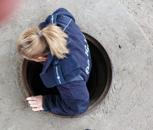Zdjęcie przedstawia umundurowaną policjantkę, która wchodzi do podziemnego przejścia przez otwarty właz.