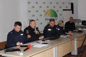 Zdjęcie przedstawia siedzących za stołem umundurowanych policjantów i umundurowanego strażnika miejskiego. Trzyma on mikrofon przy ustach.