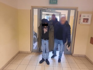 Zdjęcie przedstawia dwóch nieumundurowanych policjantów, którzy wprowadzają do budynku zatrzymanego mężczyznę. Policjant, który idzie po jego lewej stronie ubrany jest w czarna kurtkę i jasne spodnie jeansowe. Jego twarz jest zasłonięta komputerowy filtrem. Część twarzy zatrzymanego mężczyzny zasłania czarny prostokąt.