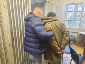 Zdjęcie przedstawia dwóch nieumundurowanych policjantów, który wprowadza do policyjnej celi zatrzymanego mężczyznę. Policjant, który idzie po jego lewej stronie ubrany jest w granatową kurtkę i jasne spodnie jeansowe. Jego twarz jest zasłonięta komputerowym filtrem. Zatrzymany mężczyzna ma na sobie długą ciemnozieloną kurtkę.