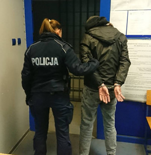 Zdjęcie przedstawia umundurowaną policjantkę, która stoi przy wyjściu z pomieszczenia z zatrzymanym mężczyzną. Oboje stoją tyłem do obiektywu aparatu. Mężczyzna ma kajdanki założone na rękach trzymanych z tyłu. Jest ubrany w szarą kurtkę i szare spodnie.