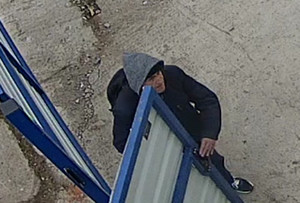 Zdjęcie przedstawia mężczyznę wchodzącego do kontenera Ma on około 45-50 lat; ubrany jest w granatową kurtkę z szarym kapturem i sportowe buty, posiada czarny plecak.