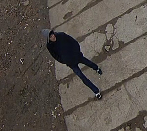 Zdjęcie przedstawia mężczyznę idącego po betonowym chodniku. Ma on około 45-50 lat; ubrany jest w granatową kurtkę z szarym kapturem, czarne spodnie i sportowe buty, posiada czarny plecak.
