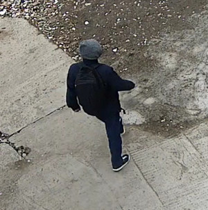 Zdjęcie przedstawia mężczyznę idącego po betonowym chodniku. Ubrany jest w granatową kurtkę z szarym kapturem, czarne spodnie i sportowe buty, posiada czarny plecak. Jest skierowany tyłem do kamery monitoringu.
