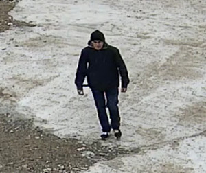 Zdjęcie przedstawia mężczyznę idącego po betonowym chodniku. Ma on około 45-50 lat; ubrany jest w granatową kurtkę z szarym kapturem, czarne spodnie i sportowe buty, posiada czarny plecak.