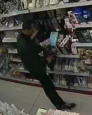 Zdjęcie przedstawia mężczyznę stojącego przy półce z asortymentem. Mężczyzna ma około 35 lat, około 185 cm wzrostu, jest szczupłej budowy ciała, ma ciemne włosy z zakolami; ubrany był w zieloną kurtkę i czarne spodnie.