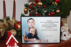 Zdjęcie przedstawia zdjęcie umieszczone w ramce, z podziękowaniami dla policjantów i pracowników Komendy Rejonowej Policji Warszawa VI. Za ramką widać świąteczny stroik oraz ubraną choinkę.