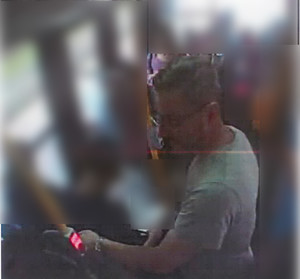 Zdjęcie przedstawia fragment sylwetki mężczyzny wewnątrz autobusu. Mężczyzna ma jasne włosy, okulary na twarzy. jest ubrany w szarą koszulkę na krótki rękaw. Lewą ręką trzyma się poręczy. Jest ustawiony lewym profilem do kamery. Tło i inni pasażerowie są rozmazani komputerowym filtrem.