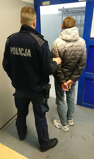 Zdjęcie przedstawia umundurowanego policjanta, który prowadzi zatrzymanego mężczyznę. Mężczyzna ten znajduje się po prawej stronie policjanta. Jest ubrany w kurtkę w kolorach jasnoszarym i ciemnoszarym. Ma na sobie niebieskie spodnie. Na rekach trzymanych z tyłu ma założona kajdanki. Obaj stoją tyłem do obiektywu aparatu. Przed nimi widać wyjście z pomieszczenia.