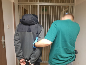 Zdjęcie przedstawia policjanta ubranego w zieloną koszulkę na krótki rękaw, który wyprowadza z pomieszczenia zatrzymanego mężczyznę. Policjant ma na dłoniach niebieskie rękawiczki ochronne. Mężczyzna ten ubrany jest w ciemną kurtkę z kapturem. Na rękach trzymanych z tyłu ma założone kajdanki. Obaj stoją tyłem do obiektywu aparatu. Przed nimi widać okratowane drzwi pomieszczenia dla osób zatrzymanych.