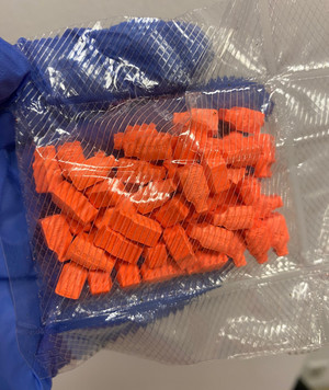 Zdjęcie przedstawia dłoń ubraną w niebieską rękawiczkę ochronną trzymającą foliowe opakowanie z pomarańczowymi tabletkami.