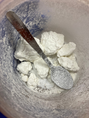 Zdjęcie przedstawia przezroczyste opakowanie w którym znajduje się biała zbrylona substancja i srebrna łyżeczka.