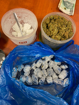 Zdjęcie przedstawia przezroczyste opakowanie w którym znajduje się biała zbrylona substancja, susz roślinny i niebieską torebkę z srebrnymi zawiniątkami. W prawym górnym rogu widać leżące na stole banknoty.