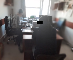 Zdjęcie przedstawia policjanta ubranego w szarą bluzę z czarnymi paskami na rękawach, który siedzi przed komputerem, prawym profilem do obiektywu aparatu. Jego twarz jest zamazana komputerowym filtrem. Tyłem do wejścia i obiektywu aparatu siedzi zatrzymany mężczyzna ubrany w ciemną kurtkę i czapkę. Część jego twarzy zasłania czarny prostokąt.