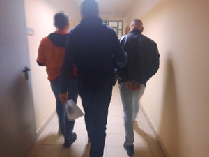 Zdjęcie przedstawia policjanta ubranego w ciemne cywilne rzeczy, który prowadzi korytarzem, po swojej prawej stronie zatrzymanego mężczyznę. Mężczyzna ten jest ubrany w czarną skórzaną kurtkę i niebieskie spodnie jeansowe. Mężczyzna na rękach trzymanych z tyłu ma założone kajdanki.