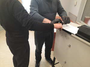 Zdjęcie przedstawia fragmenty sylwetek policjanta ubranego w granatowy sweter, czarne spodnie i czarne buty, który ma założone na rękach czarne ochronne rękawiczki. Policjant ten stoi przy stanowisku d pobierania odcisków palców i trzyma dłoń zatrzymanego mężczyzny.