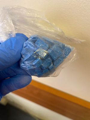 Zdjęcie przedstawia dłoń w niebieskiej ochronnej rękawiczce, która trzyma przezroczysty woreczek z niebieskimi tabletkami.