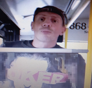 Zdjęcie przedstawia mężczyznę wewnątrz autobusu. Jest on w wieku około 40, ma 180 cm wzrostu, jest szczupłej budowy ciała. Ubrany jest w jasną koszulkę z napisem „JOKER”, czarną czapkę z daszkiem, przez ramię przewieszoną ma czarną torbę.