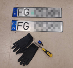 Zdjęcie przedstawia dwie tablice rejestracyjne ułożone równolegle na podłodze, obok nich widać czarne rękawiczki ochronne i śrubokręt.